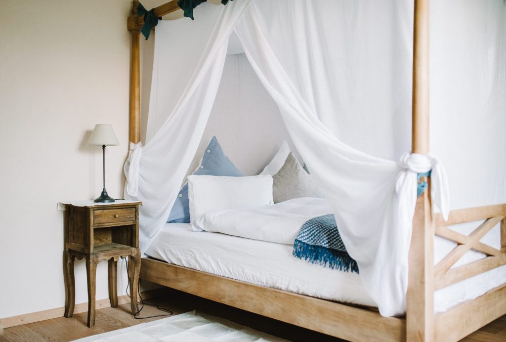 Himmelbett mit weißen Vorhängen, blauen Kissen und Holznachttisch im hellen Gästezimmer.