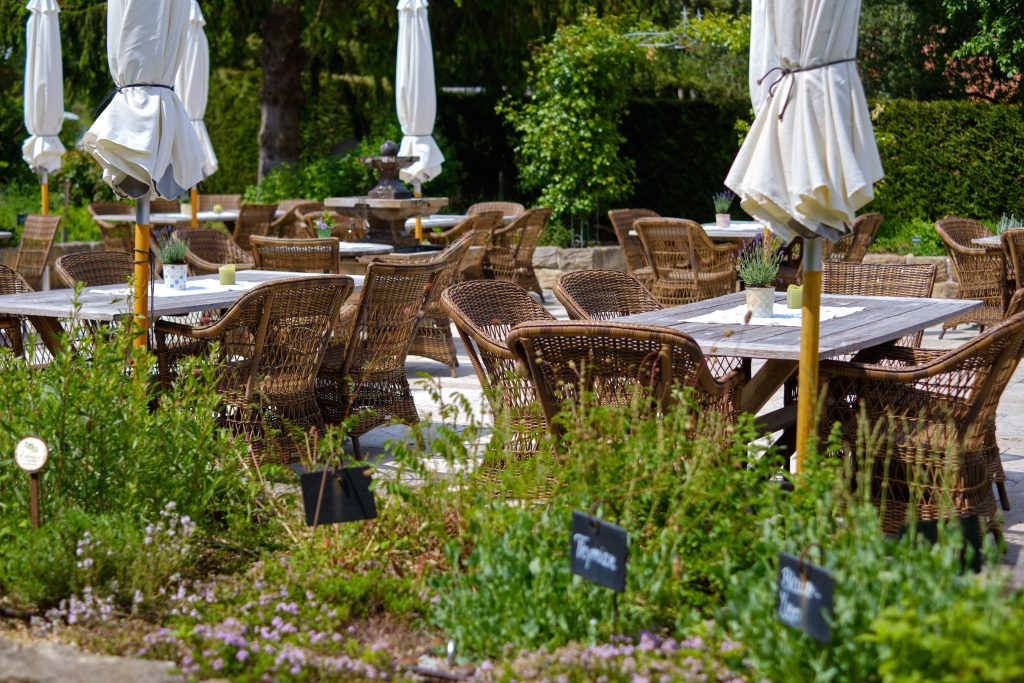 Outdoor-Bereich mit Holztischen, Rattanstühlen und geschlossenen Sonnenschirmen.